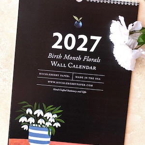 May include: A 2027 wall calendar with a black cover featuring the text 2027 Birth Month Florals WALL CALENDAR, HUCKLEBERRY PAPER, MADE IN THE USA, WWW.HUCKLEBERRYPAPER.COM, and Hand-Crafted Stationery and Gifts. It shows a blue berry and a potted flower.