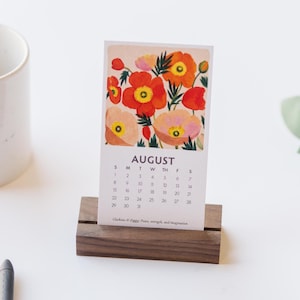 May include: A vertical paper calendar for August featuring a colorful illustration of red, orange, and peach poppies with green foliage. The calendar is held upright by a rectangular dark brown wooden stand. The text reads AUGUST and includes dates.