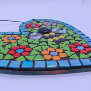 May include: A heart-shaped mosaic wall hanging with a floral design. The mosaic is made of small, colorful tiles in shades of green, blue, red, yellow, orange, and pink. The heart has a black border and a string for hanging.