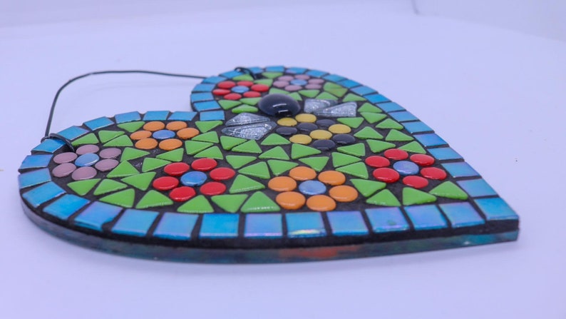 May include: A heart-shaped mosaic wall hanging with a floral design. The mosaic is made of small, colorful tiles in shades of green, blue, red, yellow, orange, and pink. The heart has a black border and a string for hanging.