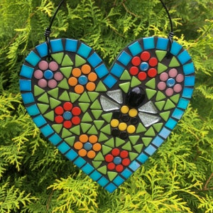 May include: A mosaic heart-shaped wall hanging with a bee design. The heart is made of small, colorful tiles in shades of blue, green, red, yellow, and black. The bee is in the center of the heart and is surrounded by flowers.