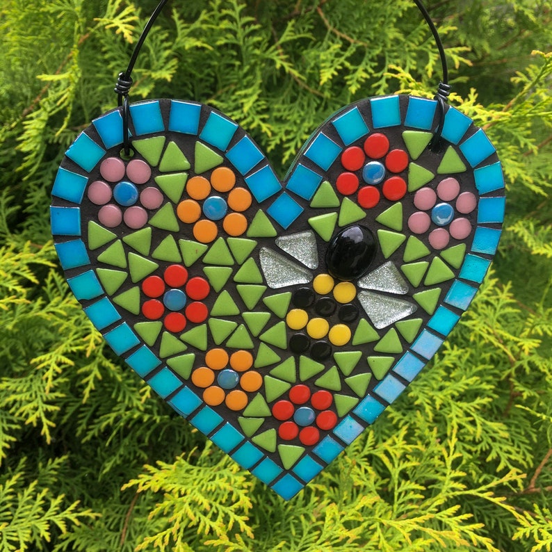 May include: A mosaic heart-shaped wall hanging with a bee design. The heart is made of small, colorful tiles in shades of blue, green, red, yellow, and black. The bee is in the center of the heart and is surrounded by flowers.