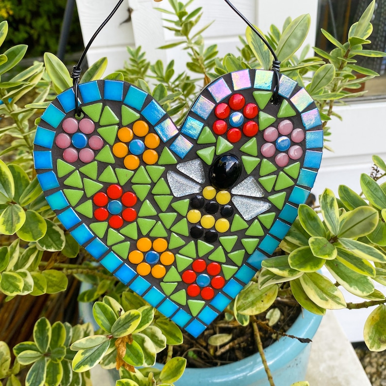 May include: A heart-shaped mosaic with a bee design. The mosaic is made of small, colorful tiles in shades of green, blue, red, yellow, and black. The bee is in the center of the heart and has a black body with yellow stripes. The heart is hanging from a black wire.