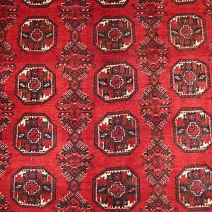 May include: A close-up of a red rug with a repeating geometric pattern. The pattern features octagonal shapes with intricate designs in black, brown, and cream. The rug has a textured appearance.