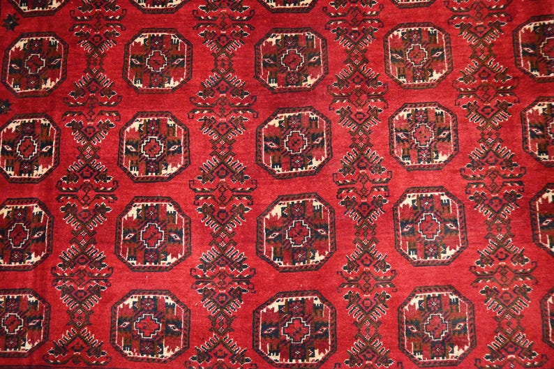May include: A close-up of a red rug with a repeating geometric pattern. The pattern features octagonal shapes with intricate designs in black, brown, and cream. The rug has a textured appearance.