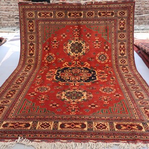May include: A large, rectangular red rug with an intricate geometric pattern. The rug features a central medallion in black and navy, surrounded by floral and geometric motifs in beige, green, and gold. The rug has a fringed edge.