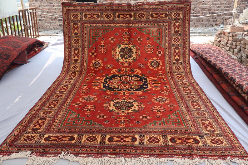 May include: A large, rectangular red rug with an intricate geometric pattern. The rug features a central medallion in black and navy, surrounded by floral and geometric motifs in beige, green, and gold. The rug has a fringed edge.