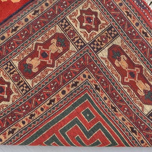 May include: Close-up of a red, beige, and green patterned rug. The rug features geometric designs and borders with repeating patterns. The rug's fringe is visible on the right side. The rug is folded over, revealing the underside.