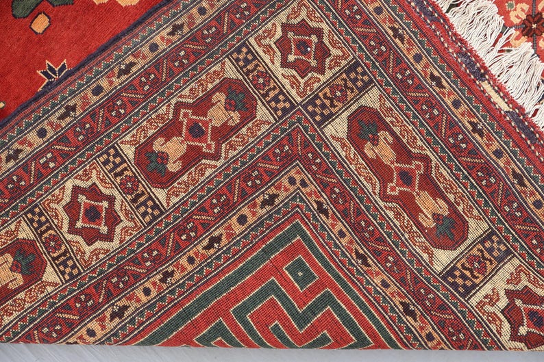 May include: Close-up of a red, beige, and green patterned rug. The rug features geometric designs and borders with repeating patterns. The rug's fringe is visible on the right side. The rug is folded over, revealing the underside.