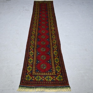 May include: A long, rectangular rug with a red central field and intricate geometric patterns. The rug features a border with blue, yellow, and green accents. The rug has a fringe on both ends.