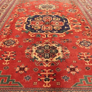 May include: A large, rectangular red rug with an intricate geometric pattern. The rug features a central medallion in navy blue, gold, and green, surrounded by repeating floral and geometric motifs in various colours. The border is a mix of red, beige, and green.
