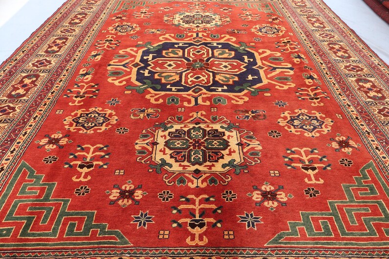 May include: A large, rectangular red rug with an intricate geometric pattern. The rug features a central medallion in navy blue, gold, and green, surrounded by repeating floral and geometric motifs in various colours. The border is a mix of red, beige, and green.