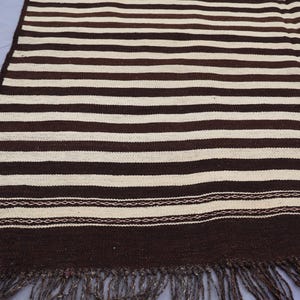 May include: A brown and white striped woven rug with fringe. The rug has a simple design with alternating stripes of brown and white.