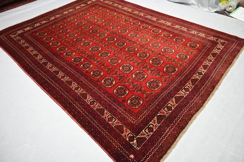 May include: A rectangular, red oriental rug with a repeating geometric pattern. The rug features a central field of repeating medallions and a detailed border in shades of red, brown, and cream. The rug's dimensions are not visible.