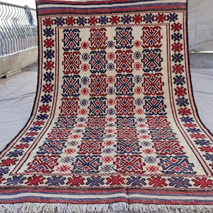 May include: A traditional rug with a geometric pattern in shades of red, blue, and white. The rug has a fringe border and is made of wool.