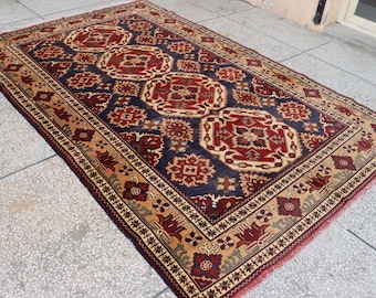 May include: A rectangular area rug featuring a dark blue central field with repeating red and cream geometric floral medallions. The border displays a tan background with intricate red and dark blue floral patterns and a fringed edge.