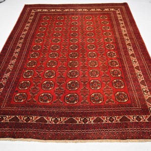May include: A rectangular, red oriental rug with a detailed geometric pattern. The rug features a central field of repeating medallions and a border with intricate designs. The color palette includes shades of red, brown, and cream.