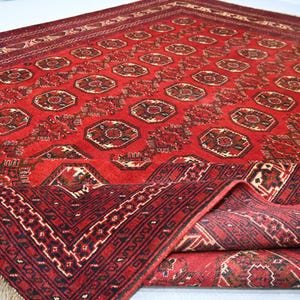 May include: A red oriental rug with a geometric pattern. The rug features a repeating design of octagons and other shapes in shades of red, brown, and cream. The rug's border has a detailed pattern and fringe.