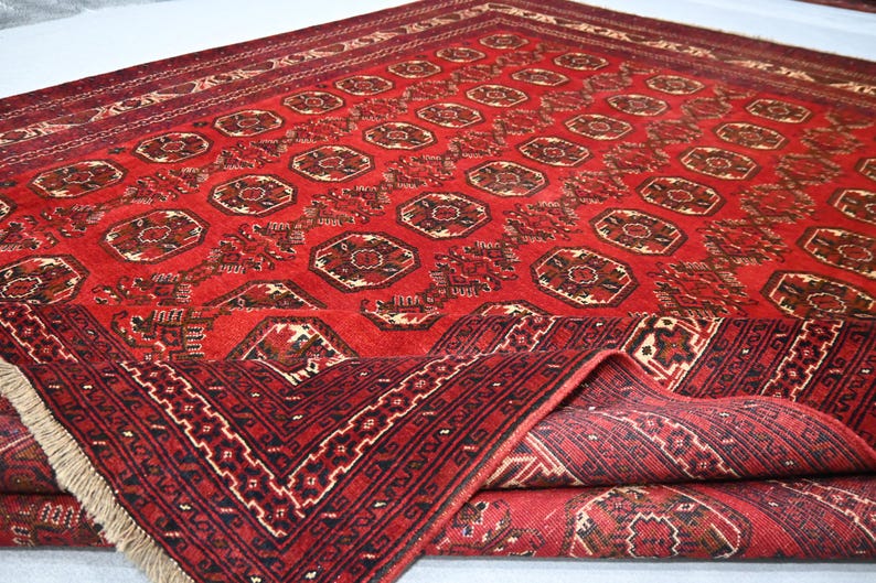 May include: A red oriental rug with a geometric pattern. The rug features a repeating design of octagons and other shapes in shades of red, brown, and cream. The rug's border has a detailed pattern and fringe.