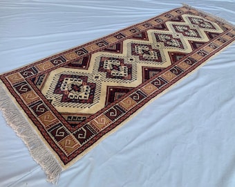 May include: A rectangular runner rug featuring a cream-colored field with a repeating pattern of dark red, navy blue, and tan geometric diamond motifs. The border displays a series of tan and dark red angular shapes, finished with white fringe.