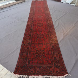 Peut inclure: Un long tapis rectangulaire rouge avec un motif géométrique brun foncé complexe. Le tapis a une frange aux deux extrémités et est présenté sur une surface grise. Le tapis est probablement un couloir.