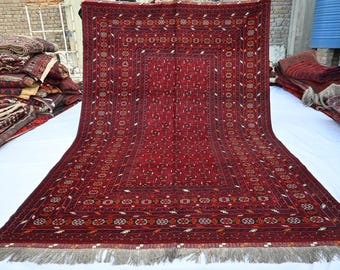 May include: A large, rectangular, red rug with an intricate geometric pattern. The rug features a central medallion design and borders with repeating floral and geometric motifs. The rug has a fringe on the bottom edge and is displayed on a white surface.