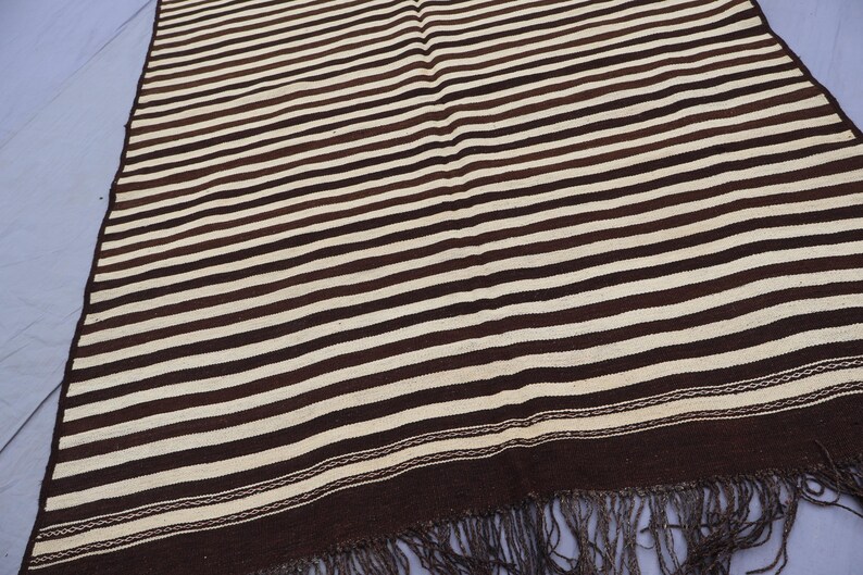 May include: A brown and white striped woven rug with fringe. The rug has a simple design with alternating brown and white stripes.