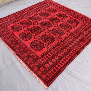 May include: A square area rug featuring a vibrant red background with repeating dark brown geometric octagonal motifs. The rug is framed by intricate dark brown borders with repeating floral and linear patterns, finished with short fringe on two sides.