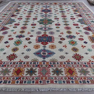 May include: A large, rectangular rug with a cream-colored background. The rug features a repeating pattern of colorful geometric shapes in red, blue, green, yellow, and pink. The border has a detailed design with fringe along the edges.