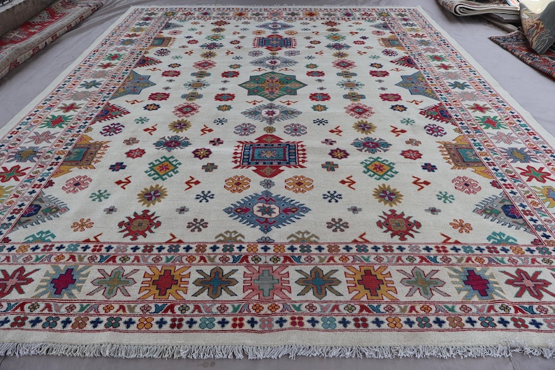 May include: A large, rectangular rug with a cream-colored background. The rug features a repeating pattern of colorful geometric shapes in red, blue, green, yellow, and pink. The border has a detailed design with fringe along the edges.