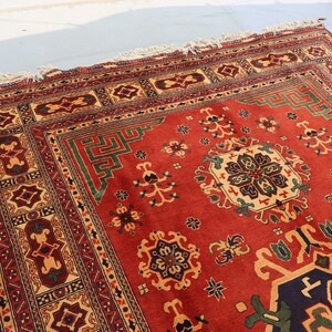 May include: A rectangular, hand-woven rug with a red field and intricate geometric patterns in beige, green, and navy blue. The rug features a detailed border and fringe along the edges, showcasing traditional craftsmanship.