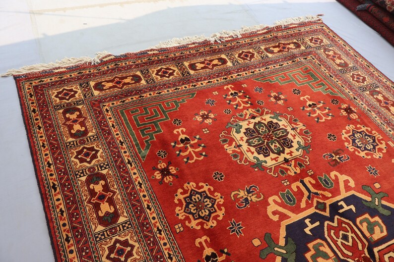 May include: A rectangular, hand-woven rug with a red field and intricate geometric patterns in beige, green, and navy blue. The rug features a detailed border and fringe along the edges, showcasing traditional craftsmanship.