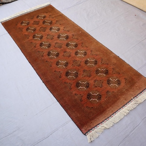 Könnte beinhalten: Ein rechteckiger, brauner Teppich mit einem sich wiederholenden Muster aus geometrischen Formen in Braun- und Cremetönen. Der Teppich hat an beiden kurzen Enden einen Fransen. Der Teppich ist wahrscheinlich aus Wolle oder einem ähnlichen Material.
