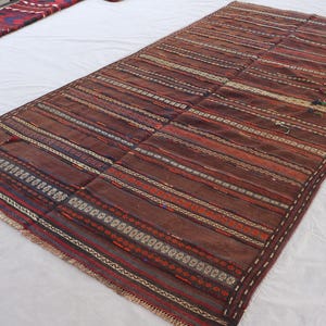 Può includere: Un tappeto rettangolare, tessuto a mano, con base marrone e strisce orizzontali nei toni del rosso, arancione e crema. Il tappeto presenta motivi intricati e un bordo sfrangiato, che mostra un design tradizionale. Adatto per soggiorno o camera da letto.