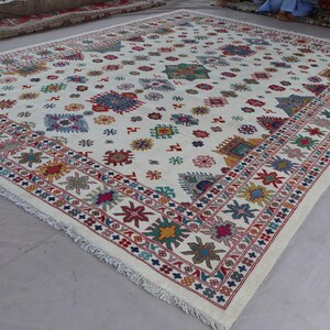 May include: A large, rectangular ivory rug with a detailed border and scattered colorful geometric patterns. The border features red, blue, and yellow floral designs. The rug's central field is adorned with various shapes in shades of blue, green, red, and yellow.