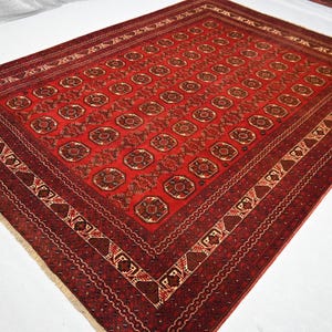 May include: A rectangular, red oriental rug with a detailed geometric pattern. The rug features a central field of repeating medallions and a border with intricate designs in shades of red, brown, and cream. The rug has a fringe on one side.
