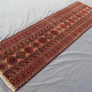 May include: A long, rectangular rug with a detailed geometric pattern in shades of red, brown, and orange. The rug has a central design with repeating circular motifs and a fringed edge. The rug is placed on a light gray surface.