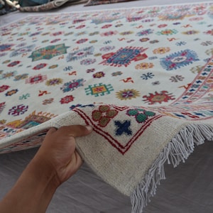 May include: Cream-colored rug with a repeating pattern of colorful geometric shapes and floral motifs. The rug has a fringed edge and a border with a repeating design. The rug is being held up to show the underside.