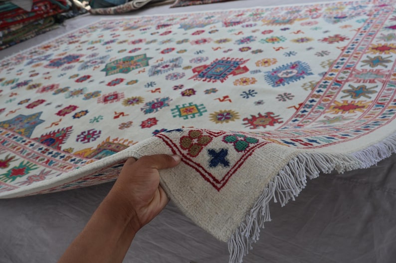 May include: Cream-colored rug with a repeating pattern of colorful geometric shapes and floral motifs. The rug has a fringed edge and a border with a repeating design. The rug is being held up to show the underside.