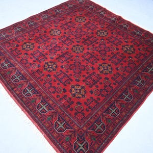 May include: A square, red rug with an intricate geometric pattern. The rug features a central design with repeating shapes in shades of red, blue, and gold. The border has a detailed pattern with fringe along the edges.