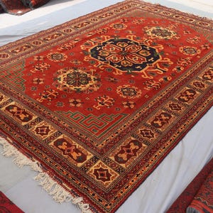 May include: A rectangular red rug with an intricate geometric pattern. The rug features a central medallion in navy and gold, surrounded by floral and geometric motifs. The border is detailed with repeating patterns and fringe.