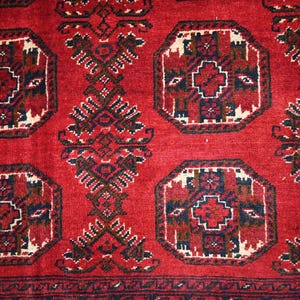 May include: A close-up of a red rug with an intricate geometric pattern. The design features repeating octagonal shapes with a central cross motif, framed by a white border. Dark brown and navy blue accents add contrast to the red background.