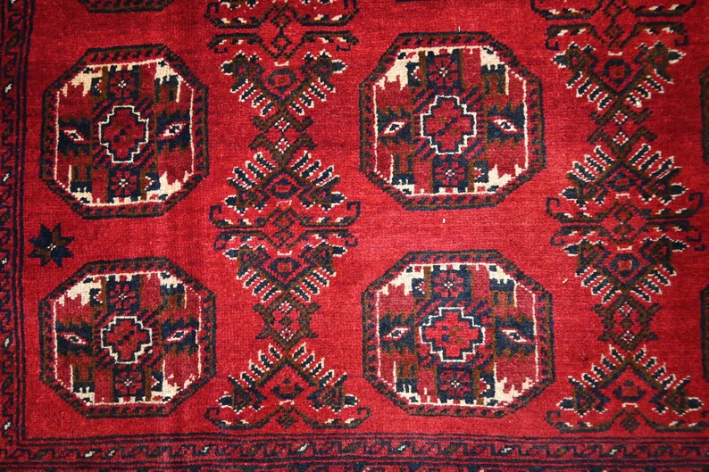 May include: A close-up of a red rug with an intricate geometric pattern. The design features repeating octagonal shapes with a central cross motif, framed by a white border. Dark brown and navy blue accents add contrast to the red background.