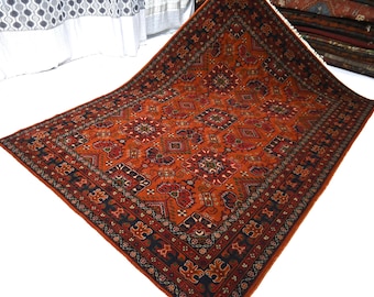 Könnte beinhalten: Ein rechteckiger Teppich mit einem leuchtend rostorangen Hintergrund und komplizierten geometrischen Mustern in Marineblau, Creme und Dunkelrot. Der Teppich hat eine breite, detaillierte Bordüre mit wiederkehrenden Motiven und kurzen weißen Fransen.