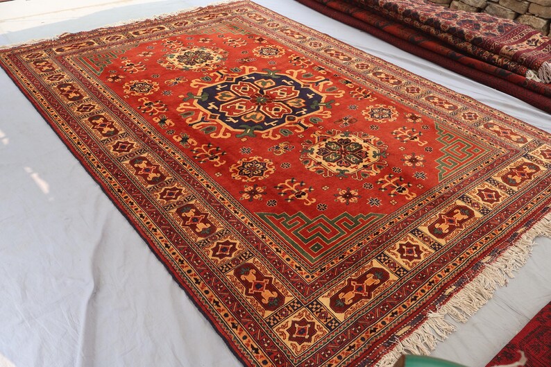 May include: A red oriental rug with a central medallion design and a detailed border. The rug has intricate geometric patterns in blue, green, and gold. The rug has a fringed edge.