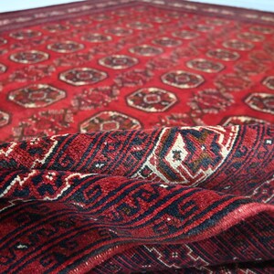 May include: A red oriental rug with a detailed geometric pattern. The rug features a repeating design of medallions and floral motifs in shades of red, black, and cream. The rug is partially rolled up, revealing the intricate weave.