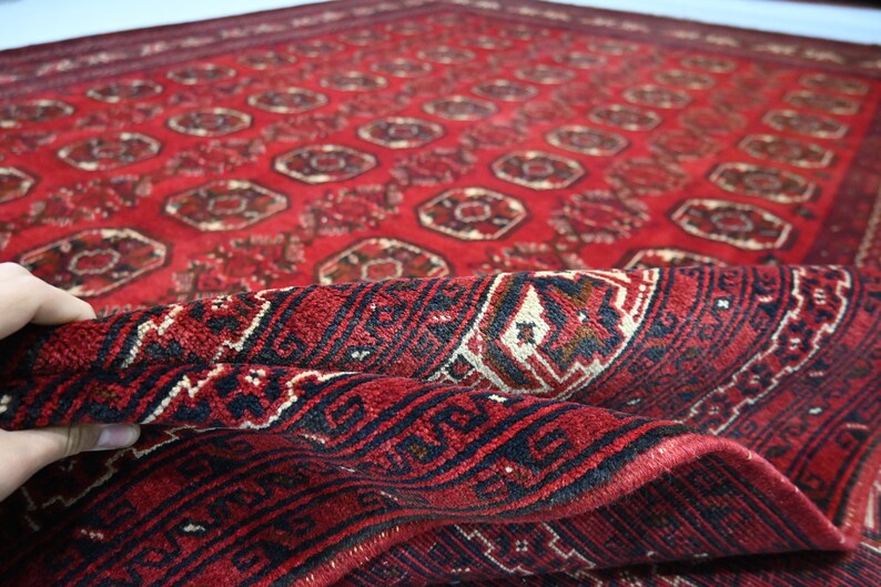 May include: A red oriental rug with a detailed geometric pattern. The rug features a repeating design of medallions and floral motifs in shades of red, black, and cream. The rug is partially rolled up, revealing the intricate weave.