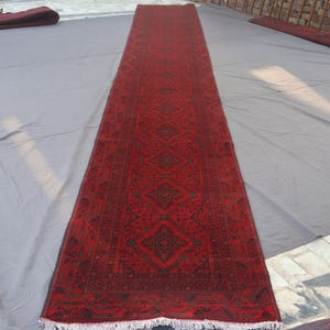 Puede incluir: Una alfombra roja rectangular y larga con un patrón geométrico. La alfombra presenta un diseño de diamantes repetidos en un tono más oscuro de rojo y negro. La alfombra tiene flecos blancos en ambos extremos. La alfombra mide aproximadamente 3 metros de largo.