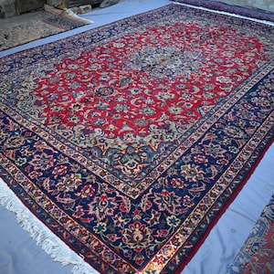 May include: A large, rectangular red rug with an intricate floral pattern. The rug has a navy blue border with floral designs and white fringe along the edges. Additional rugs are visible in the background.