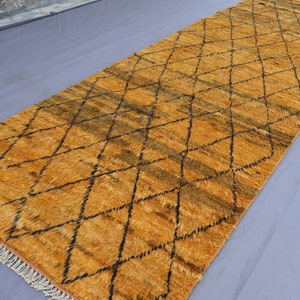 Puede incluir: Una alfombra tejida a mano con un patrón de diamantes en tonos marrones y amarillos. La alfombra tiene un borde con flecos.